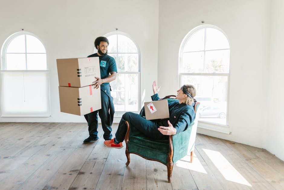 A man with dark curly hair is kneeling on a wooden floor inside a room, surrounded by several large cardboard moving boxes of various sizes, some labeled with room and size details. He is wearing a te
