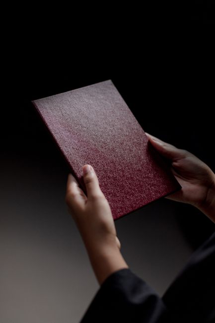 Hands holding a closed Victorian wardrobe made of dark wood with a textured maroon and grey fabric cover, positioned indoors. The wardrobe is part of a house removal process, viewed from a slightly el