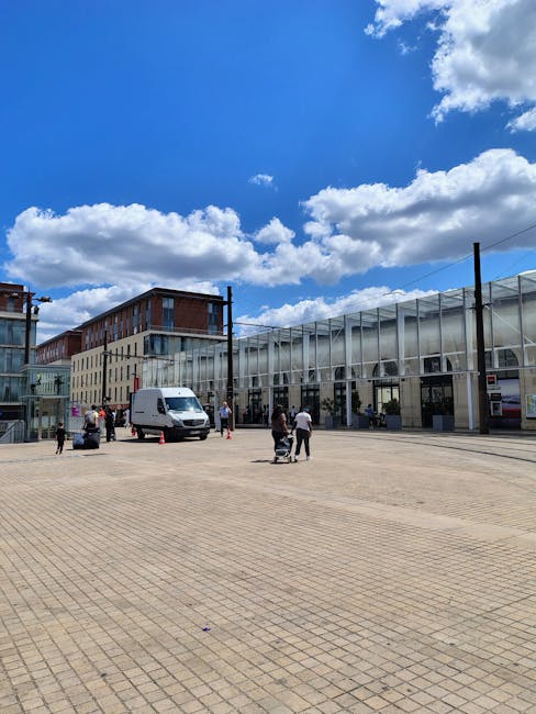 A clear day scene outside a modern commercial building at Copeland Park with a large paved area in the foreground. Several people are visible, some pushing carts or carrying items, indicating a moving