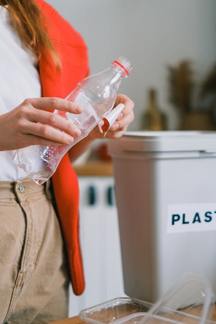 A person wearing a plain white T-shirt is holding a large green recycling bin with a white recycling symbol on the front. The individual’s face is not visible, and they are standing against a solid 
