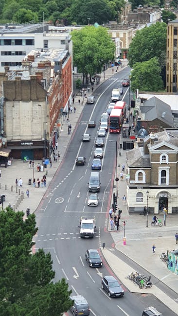 An aerial view of a busy urban street in Peckham, showing a line of parked cars along the curb and moving vehicles, including a red double-decker bus and several smaller cars, traveling in the same direction. The street is bordered by mixed-use buildings, including a brick structure on the left with a storefront and beige residential houses on the right. Sidewalks on both sides are populated with pedestrians, some walking or waiting at crossings, and a few bicycles parked near the pavement. Green trees line the street, providing shade and contrast to the cityscape. Visible on the pavement are road markings and pedestrian crossings, with some people crossing the street. This scene depicts typical urban traffic and pedestrian activity, relevant to home relocations and furniture transport projects managed by companies such as Man and Van Peckham, highlighting the environment where moving and packing logistics often take place.