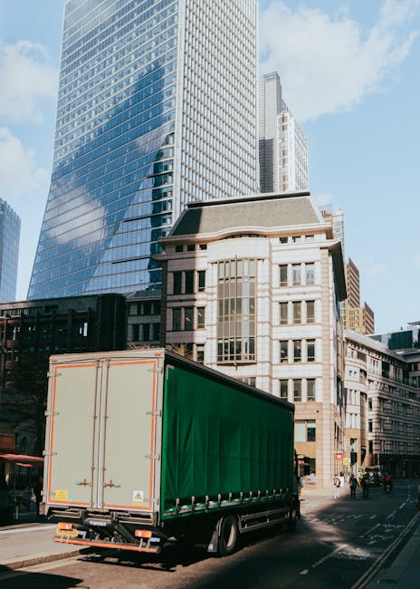 A large delivery truck with a green cargo cover is parked on a city street, adjacent to modern and historic buildings, including a tall glass skyscraper reflecting the sky and surrounding structures. The truck is positioned near the sidewalk, with its rear doors closed and secured, ready for a home relocation or furniture transport. The surrounding environment features pedestrians walking on the pavement, and a few cyclists, indicating an active urban area. The scene is illuminated by natural daylight, highlighting the reflective glass surfaces and the architectural details of the nearby buildings. The image captures the loading or unloading process typical of professional removals services by Man and Van Peckham, where furniture, boxes, and packing materials might be loaded onto or off the vehicle as part of the moving logistics within central London.