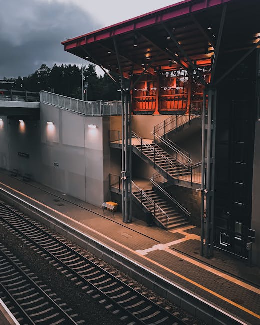 A narrow, elevated pathway with a grey asphalt surface runs alongside a train station platform, enclosed by a blue metal railing with yellow handrails. On the left side, there is dense greenery with t